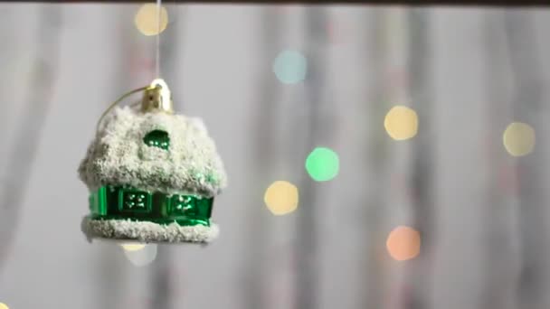 Рождественская елка игрушка в виде зеленого домой.Красивая елка игрушка висит на фоне разноцветных рождественских огней и слегка движется. Close-up .Christmas tree toy in form house.  - Кадры, видео
