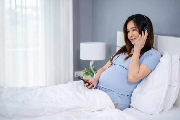 femme enceinte utilisant un smartphone pour écouter de la musique grâce à des écouteurs sur un lit - Photo, image