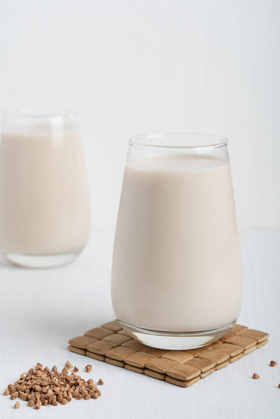 Gluten-free and lactose-free healthy buckwheat milk full of vitamins, minerals and antioxidants served in tall drinking glass on wooden mat with raw grain ingredient on white background. Vertical - Foto, Imagen