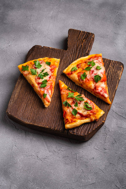 Tranches de pizza chaudes avec fromage mozzarella, jambon, tomate et persil sur planche à découper en bois marron, fond en béton de pierre, vue d'angle - Photo, image