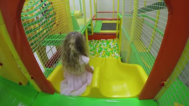 Active girl in pink dress slide down into colorful balls pool at playground - Footage, Video
