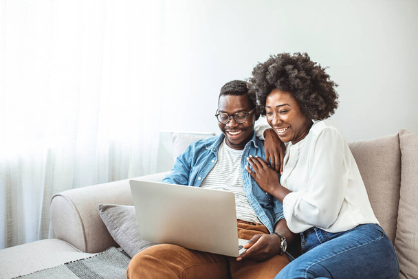 Young black couple sit on the sofa using laptop. Black couple using laptop at home look at each other. Overjoyed african American couple sit relax on cozy couch look at laptop screen triumph winning lottery online - Zdjęcie, obraz