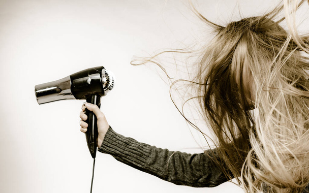 Blonde woman styling her very long hair. Teenage girl with blowing messy hair holding hairdryer. - Фото, изображение