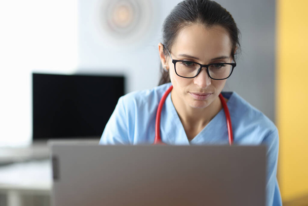Młoda kobieta lekarz w okularach siedzi na laptopie w gabinecie kliniki - Zdjęcie, obraz