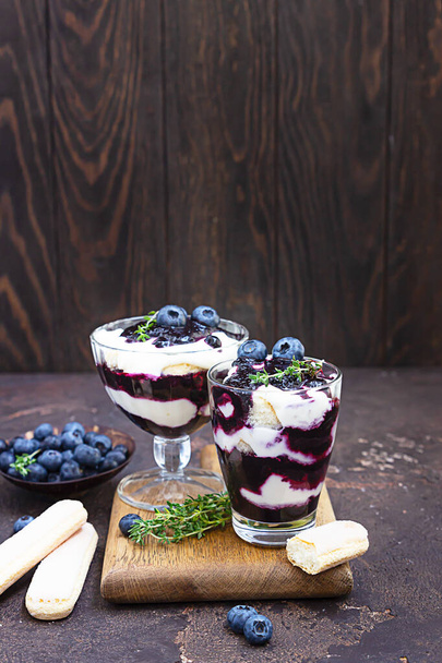 Dessert from mascarpone or ricotta cheese, blueberries and biscuit in glass on a dark concrete background. No baked cheesecake or tiramisu. - Foto, immagini