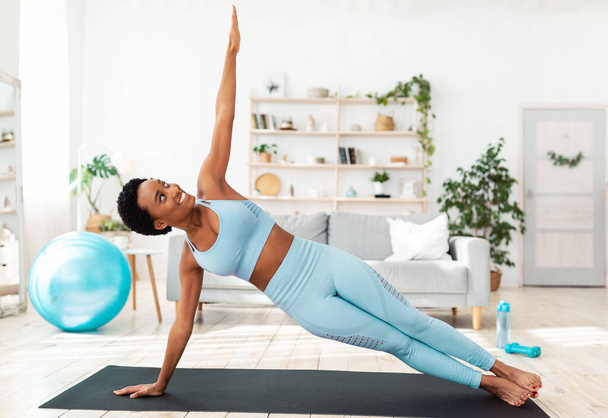 Home fitness concept. Determined black woman working out on sports mat indoors, doing side plank exercise - Foto, imagen