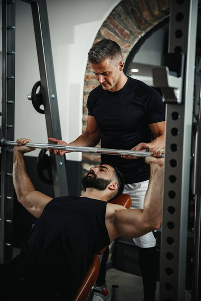Aufnahme eines muskulösen Kerls in Sportbekleidung, der mit Personal Trainer im Fitnessstudio trainiert. Er pumpt bei hartem Training die Brustmuskulatur auf. - Foto, Bild