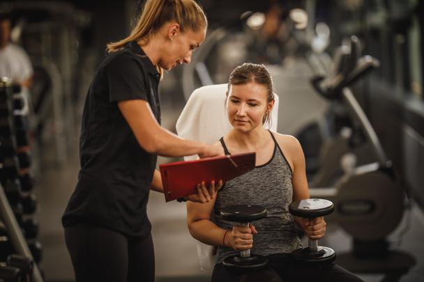 Shot of a muscular young woman in sportswear working out with female personal trainer at the gym. She is pumping up her muscule with dumbbell. - Photo, Image