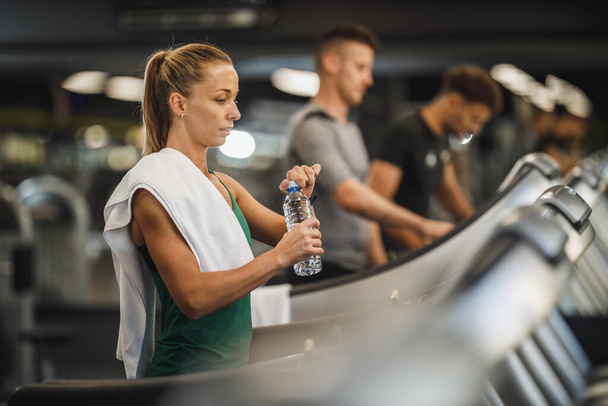 Shot of a young woman walking on a treadmill at the gym. She is getting to drink water. - Φωτογραφία, εικόνα