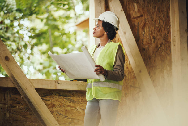 Prise de vue d'une architecte africaine vérifiant les plans sur le chantier d'une nouvelle maison en bois. Elle porte des vêtements de travail protecteurs et un casque blanc. - Photo, image