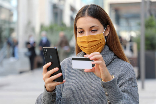 Портрет молодої красивої жінки в захисній масці охолоджується і робить оплату через кредитну картку і додаток для смартфона на вулиці
 - Фото, зображення