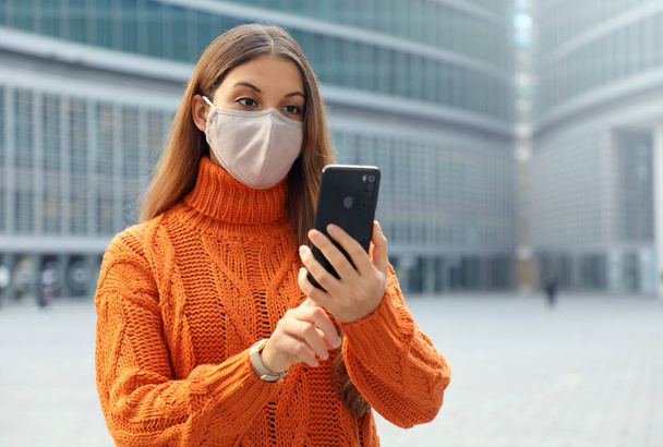 Νεαρή γυναίκα φοράει μάσκα προσώπου χρησιμοποιώντας την εφαρμογή smartphone στο σύγχρονο δρόμο της πόλης. Αντιγραφή χώρου. - Φωτογραφία, εικόνα