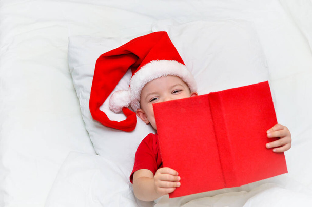 El pequeño Santa lee un libro rojo y yace en su cama bajo una manta blanca. - Foto, imagen