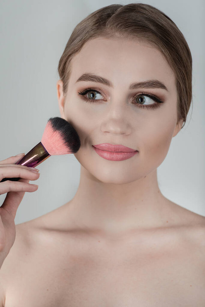 Beautiful young woman applying cosmetic powder on her face with tassel, skin care concept. photo composition of brunette girl on gray background - Photo, Image