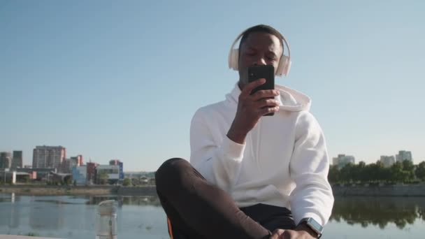Medium close up of young african american man with headphones sitting on embankment scrolling on his smartphone with river in background - Footage, Video