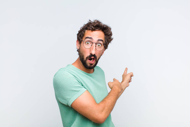 young bearded man feeling shocked and surprised, pointing to copy space on the side with amazed, open-mouthed look - Photo, image