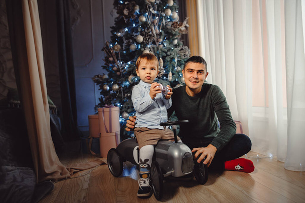 Ο μπαμπάς και ο γιος του παίζουν κάτω από το χριστουγεννιάτικο δέντρο. Δώρα κάτω από το χριστουγεννιάτικο δέντρο. Χριστουγεννιάτικη ιδέα, δώρα, αναμνήσεις της παιδικής ηλικίας. - Φωτογραφία, εικόνα