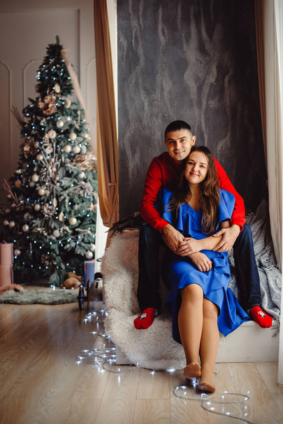 Pareja joven, hombre y mujer posando bajo el árbol de Navidad con regalos en el interior. Concepto de Navidad, regalos, recuerdos de la infancia. - Foto, imagen