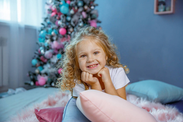 Ένα κοριτσάκι κοντά στο χριστουγεννιάτικο δέντρο είναι ξαπλωμένο στο κρεβάτι.   - Φωτογραφία, εικόνα
