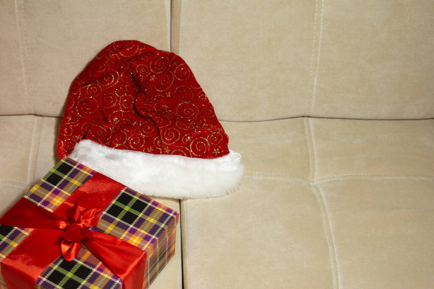 copy space. close up. horizontal. festive red santa claus hat, festive black checkered gift box wrapped in a red ribbon are on the light cream sofa. minimalism. holiday symbols. New Year. Christmas. - Foto, Imagem
