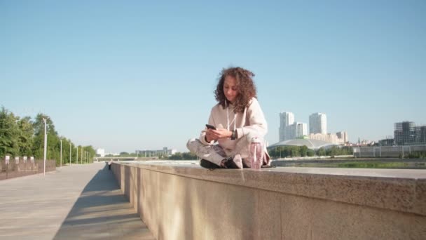 Full shot of smiling caucasian woman with curly hair in sporty hoodie and leggings sitting at embankment checking her phone recreating after jogging - Footage, Video