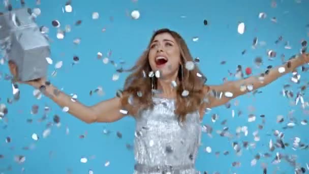 Cámara lenta de mujer feliz saltando con regalo cerca de confeti sobre fondo azul  - Imágenes, Vídeo