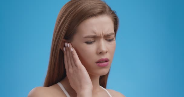 Mujer joven trastornada sosteniendo el oído doloroso, sintiendo repentinamente dolor fuerte, problema de salud, fondo azul, primer plano - Metraje, vídeo