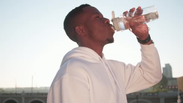 Low-angle close up of young african american sportsman drinking water from sports water bottle looking straight ahead after hard outdoor workout at dawn - Footage, Video