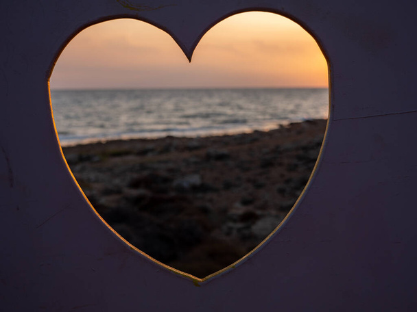 心臓の形の穴を通って表示されます。人々は海岸を歩いてビデオを作っています。愛、ケア、バレンタインのロマンチックなコンセプト. - 写真・画像