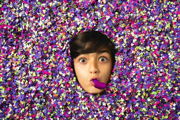Удивлённое лицо мальчика, дующего в рог вечеринки, окружённого цветными конфетти. - Фото, изображение