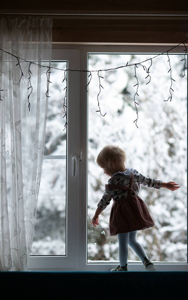 Toddler lány áll az ablakon díszített karácsonyra és nézi az első hó. Újévi ünnepek. Függőleges formátum - Fotó, kép