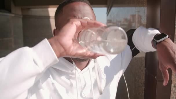Primo piano di bel corridore afro-americano maschio con cuffie e smartwatch acqua potabile dalla bottiglia sportiva dopo il jogging all'aperto - Filmati, video