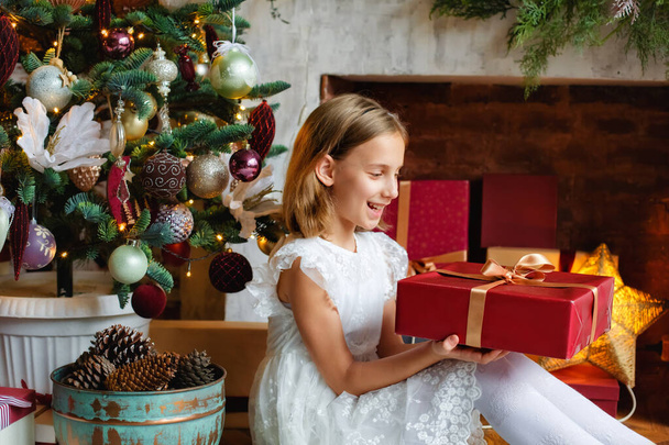 Navidad, vacaciones y concepto de la infancia-una chica sonriente con una caja de regalo en casa cerca de un árbol de Navidad decorado. - Foto, Imagen