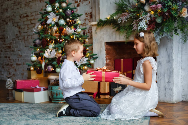 Çocuklar Noel hediyelerini açın. Noel ağacının altında hediye kutularıyla çocuklar. Geleneksel şöminesi olan dekore edilmiş oturma odası. Evde sıcak bir kış günü.. - Fotoğraf, Görsel