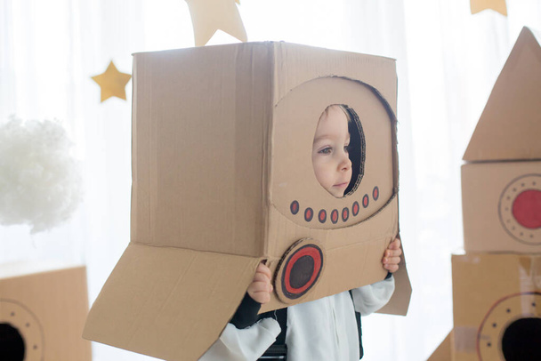 Sweet toddler boy, dressed as an astronaut, playing at home with cardboard rocket and handmade helmet from box - Foto, imagen