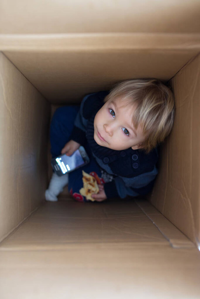 Sweet toddler child, cute blond boy, hiding in cardboard box, looking out from a hole, smiling - Φωτογραφία, εικόνα