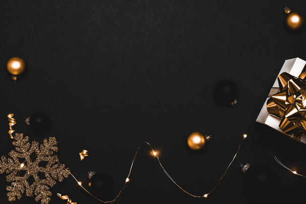 Vrolijk kerstfeest. Wit cadeau met gouden strik, gouden ballen en sprankelende lichtslinger in kerstversiering op donkere achtergrond voor wenskaart. Kerstmis, winter, nieuwjaarsconcept - Foto, afbeelding