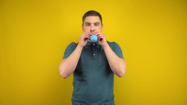 A young man inflates a blue balloon with his mouth on a yellow background. Man in a green polo. - Footage, Video