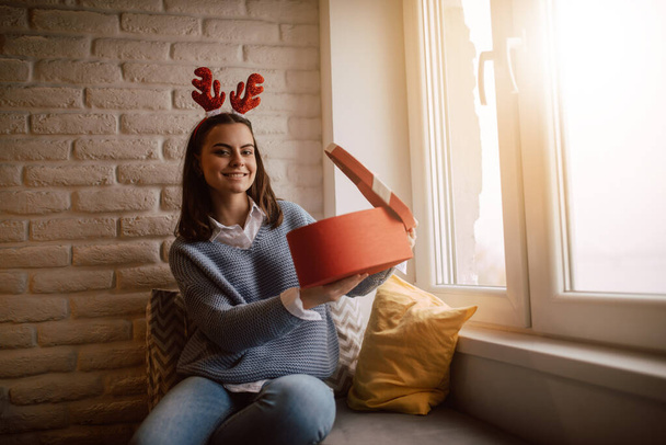 Ein fröhliches junges kaukasisches Mädchen mit einer Hirschohren-Requisite auf dem Kopf zeigt eine rote Geschenkschachtel mit Schleife. Neujahr und Weihnachten zu Hause feiern - Foto, Bild
