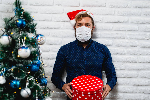 Egy szakállas férfi arcmaszkban egy karácsonyfa mellett, egy karácsonyi ajándékdobozzal a kezében. Újév és Karácsony a karanténban a világjárvány idején COVID - 19 koronavírus - Fotó, kép