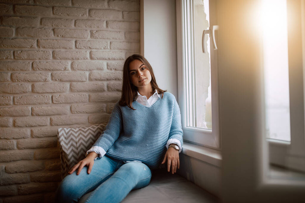 Retrato de uma jovem bela adolescente caucasiana sentada em um sofá junto à janela na sala de estar e olhando para a câmera. Relaxe o tempo na casa - Foto, Imagem