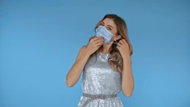 Mujer alegre quitándose la máscara médica aislado en azul  - Metraje, vídeo