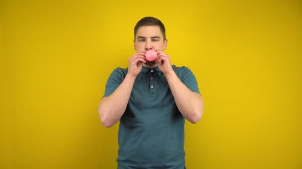 A young man inflates a pink balloon with his mouth on a yellow background. Man in a green polo. - Footage, Video
