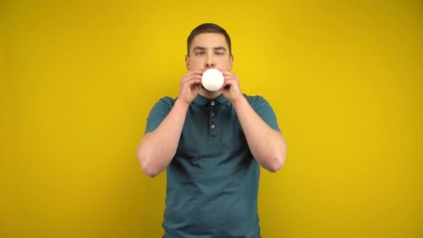 Un jeune homme gonfle un ballon blanc avec sa bouche sur un fond jaune. Homme dans un polo vert. - Séquence, vidéo
