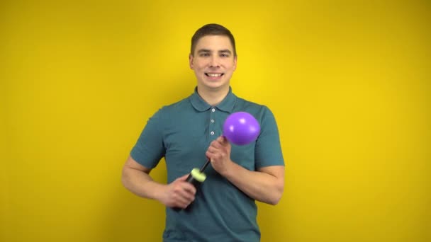 A young man inflates a purple balloon with a pump on a yellow background. Man in a green polo. - Footage, Video