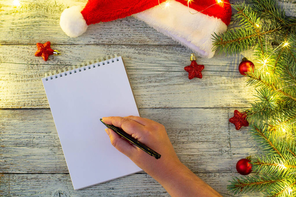 χέρι της γυναίκας γραφής επιστολή σε χαρτί σημειωματάριο με χριστουγεννιάτικες διακοσμήσεις και bokeh σε λευκό ξύλινο φόντο - Φωτογραφία, εικόνα