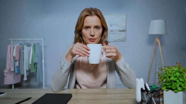 focused woman looking at camera and drinking tea during skype call at home - Materiał filmowy, wideo