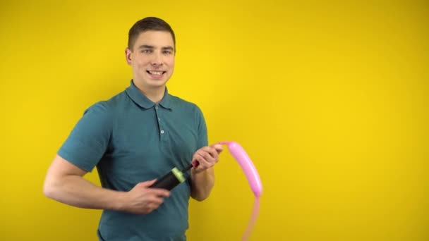 若い男が黄色の背景にポンプで長いピンクの風船を膨らませています。緑のポロの男. - 映像、動画