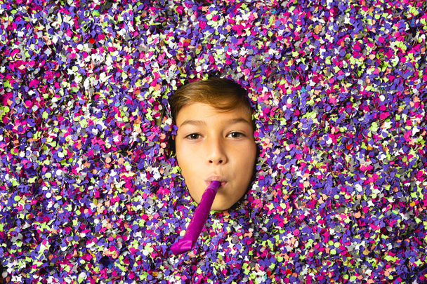 Joven cara de niño tocando un cuerno de fiesta y estar rodeado de confeti de colores - Foto, imagen