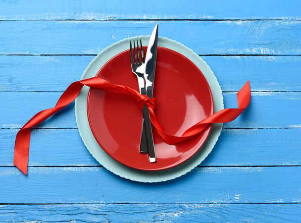 round plate and metal fork and knife tied with a red silk ribbon, blue wooden background, top view - Photo, Image
